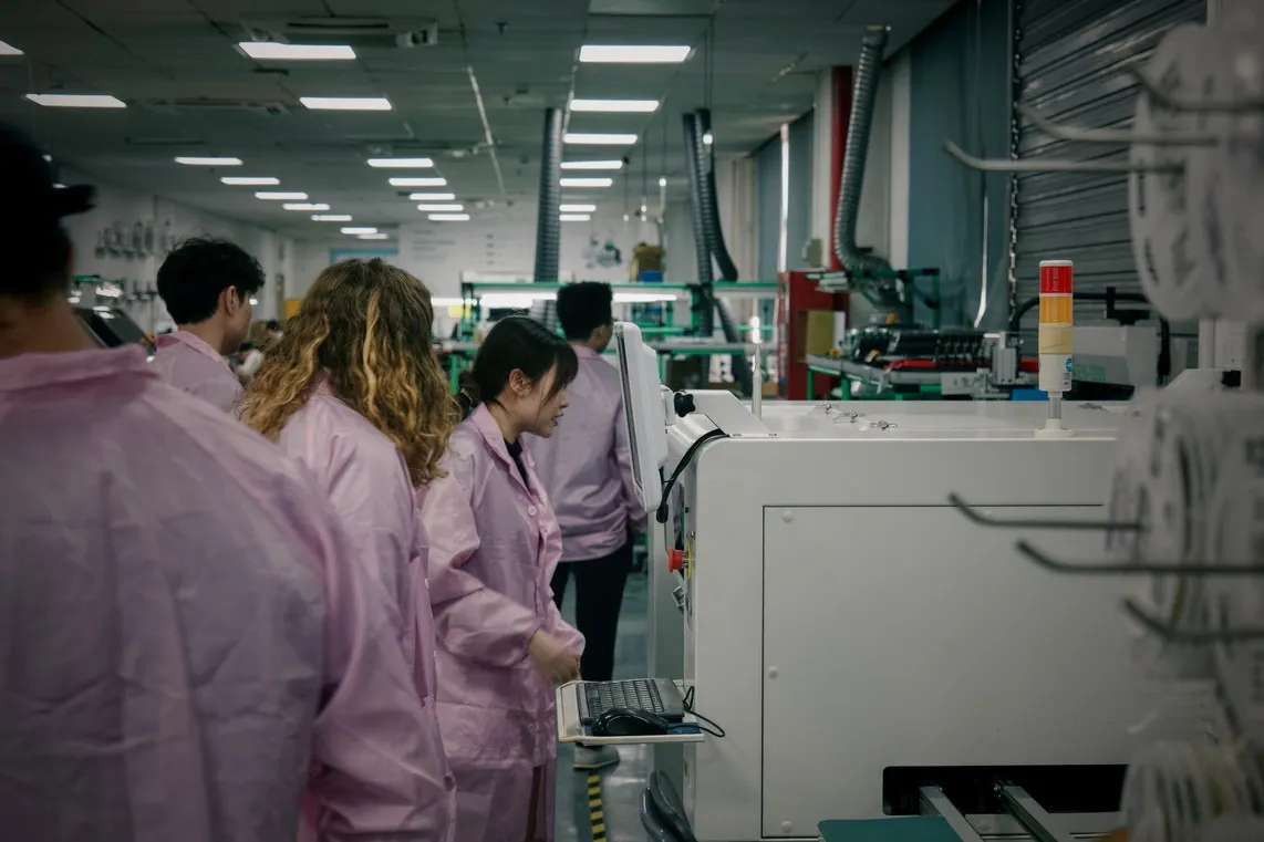 A group of visiting founders in pink ESD smocks walking past an SMT line in a Shenzhen electronics factory
